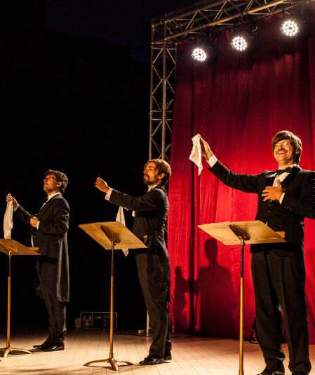 Affiche du spectacle. Photo de trois chanteurs devant un rideau de fond de scène rouge et des projecteurs, en train de chanter.