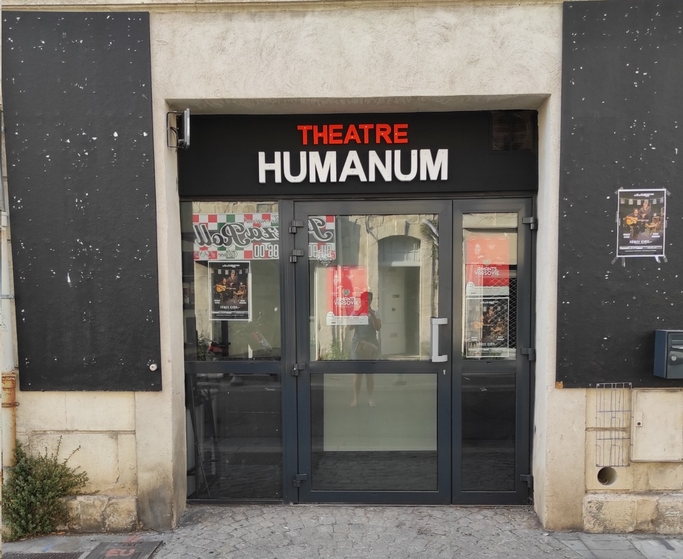 Photo de la façade du théâtre. Deux panneaux verticaux noir servant à mettre les affiches des spectacles encadrent l’entrée du théâtre. La porte est vitrée, et le nom du théâtre apparaît en couleurs au-dessus.
