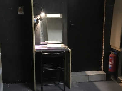 L’espace loge qui est une table en bois avec un miroir contre le mur, une lampe et une chaise noire. À sa droite on peut voir la porte des toilettes.